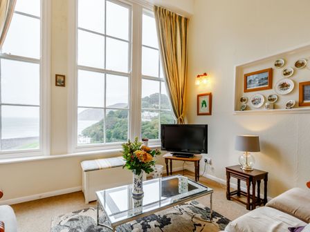 A living room with large windows overlooking a coastal landscape with hills a TV on a wooden stand a glass coffee table with flowers a lamp on a side table and framed pictures with plates on the wall at Clooneavin Apartment 2 in Lynmouth