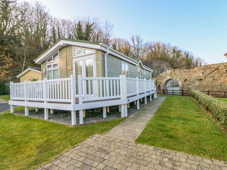 A wooden cabin with white railings on a raised platform near a stone archway and trees at The Hideaway - Heritage Park in Stepaside near Kilgetty