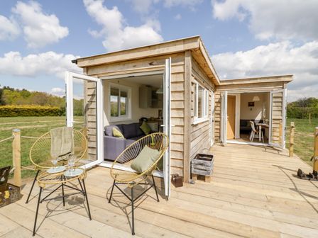An outdoor wooden deck with two chairs and a table with glasses outside a wooden lodge with open doors showing a living room and dining area at Malvern View Lodge in Wadborough near Pershore