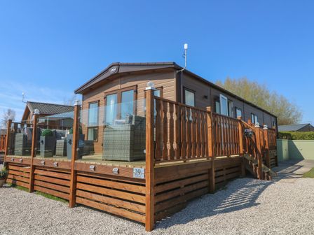 A wooden lodge with a glass and wood railing deck and outdoor wicker furniture at Lancaster Lodge in South Lakeland Leisure Village