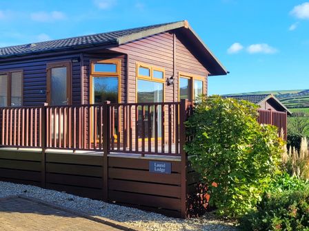 A wooden lodge with a deck and railing surrounded by bushes at Laurel Lodge Bossiney Bay Holiday Park near Tintagel