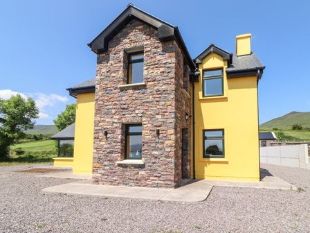 A house with yellow walls and stone accents on a gravel lot with hills and trees in the background at Lack Cottage Annascaul near Camp County Kerry