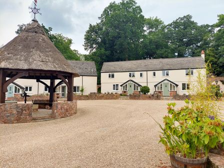 A courtyard with a thatched gazebo and two white cottages with green doors surrounded by trees at Pumphouse Cottage in Uffculme