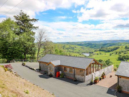 A wooden house with a gray roof on a paved road with green hills and a lake in the background at The Larches Van near Llanidloes