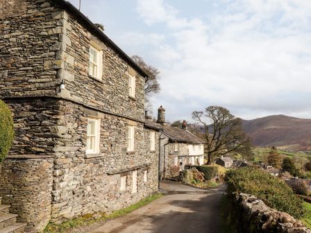 A stone house next to a road with trees and hills in the background at Rose Cottage At Troutbeck in Troutbeck