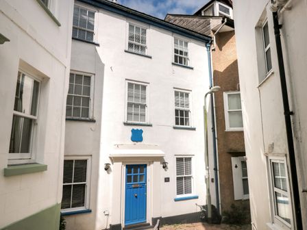 A white building with a blue door and windows in a narrow alley at Grants Cottage in Dartmouth