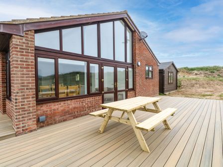 A wooden picnic table on a deck outside a brick house with large windows and a wooden shed in the background at Windyridge in Eccles-on-Sea near Sea Palling