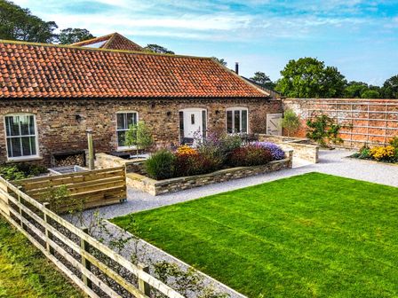A garden with a lawn flower beds gravel paths and a brick building with a tiled roof at Flittermouse Barn in Hornsea