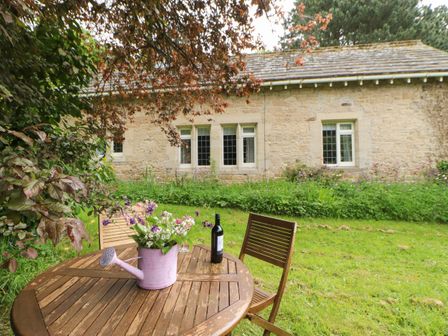 A garden with wooden table and chairs a watering can with flowers and a bottle of wine on the table in front of a stone house at The Coach House in Shepherd's Dene near Corbridge