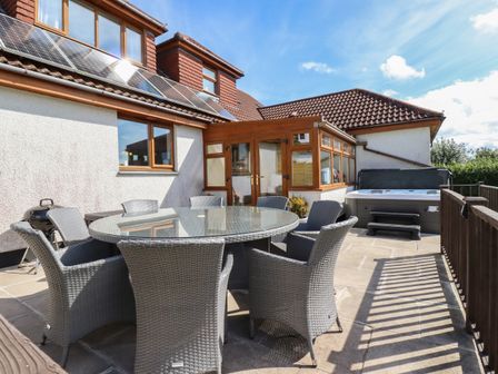 An outdoor patio with a round glass table and six wicker chairs next to a white house with solar panels and a hot tub at Meadowside in Goonhavern