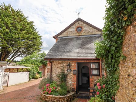 A stone and brick cottage with a garden featuring pink flowers and greenery and a small table and chair outside the entrance at Coach Cottage in Colyton