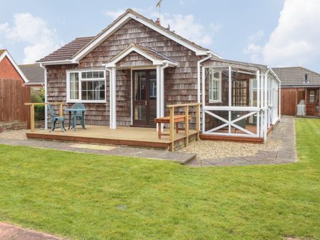 A wooden house with a small porch table and chairs and a glass-enclosed patio in a grassy yard at Doves Nest in Martham