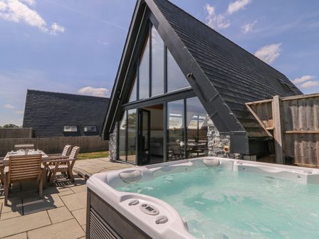An outdoor patio with a hot tub and wooden dining table with chairs next to a house with large glass windows at Hill Radnor - The Sheepfold in Llanyre near Llandrindod Wells