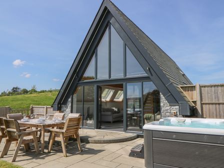 An outdoor patio with a dining table and chairs and a hot tub in front of an A-frame building with large glass windows at Brecknock Cheviot - The Sheepfold in Llanyre near Llandrindod Wells