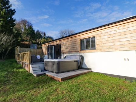 A wooden exterior house with a deck area with seating and a hot tub on a grassy lawn at The Linhay in Cadeleigh near Tiverton
