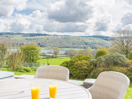 A patio with a round wooden table and wicker chairs overlooking a green landscape with trees and a lake at Cedar Wood Coniston in Coniston