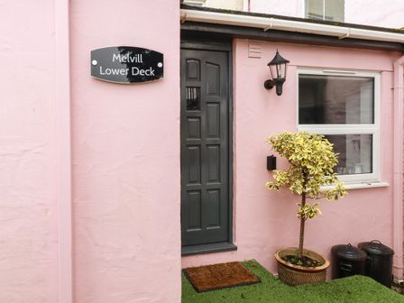 The entrance to a building with a black door pink walls a potted plant two trash bins and a sign that reads Melvill Lower Deck in Falmouth