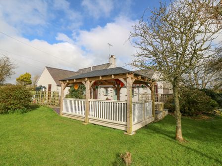 A wooden gazebo in a garden with a tree and a house in the background at Ty Gwennol in Llanfaethlu
