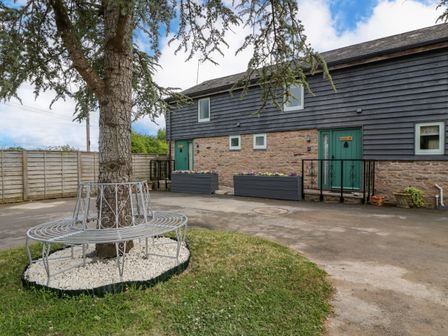 A courtyard with a tree surrounded by a metal circular bench and a building with green doors at Beekeeper's Cottage in Pembridge