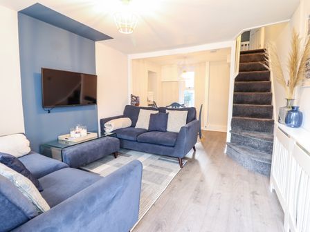 A living room with blue sofas a wall-mounted TV a glass coffee table a carpet and a staircase at Driftwood Terrace in Porthmadog