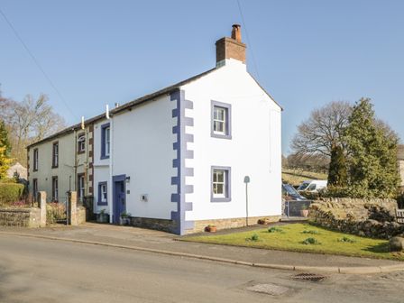 A street view of a white two-story house with blue window trims a chimney and a stone wall beside it in Caldbeck