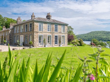 A large stone house with multiple chimneys surrounded by grass and trees near a body of water at Silverholme Graythwaite near Hawkshead