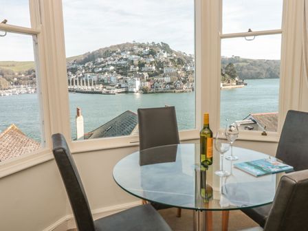 A dining area with a round glass table and four chairs next to windows overlooking a river and hillside town at 4 Dartview in Dartmouth