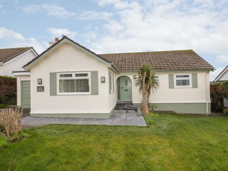 The front view of a single-story house with a tiled roof a lawn a paved path and green shutters at Hardy Cottage in Benllech