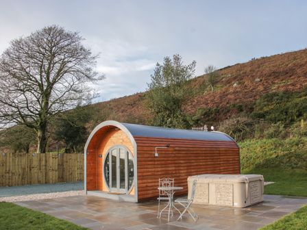A wooden pod cabin with a round glass door a small patio table and chairs and a hot tub outside on a paved area at Stapeley Pod in White Grit near Chirbury
