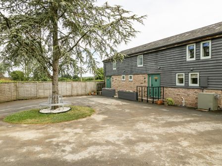 An outdoor area with a tree surrounded by a circular bench and a building with green doors at Bluebell Cottage in Pembridge