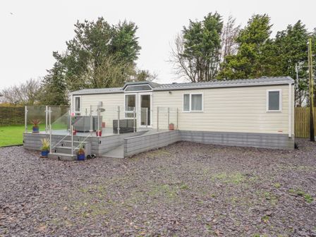 A mobile home with a deck and glass railing surrounded by trees at Banadlen Lodge in Brynteg near Benllech
