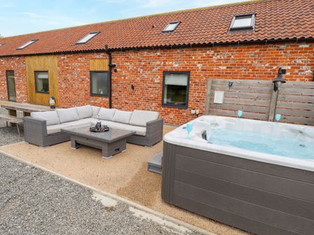 An outdoor patio area with a grey cushioned corner sofa and glass coffee table beside a hot tub and a red brick building at Pheasant's Roost in Lelley near Burton Pidsea