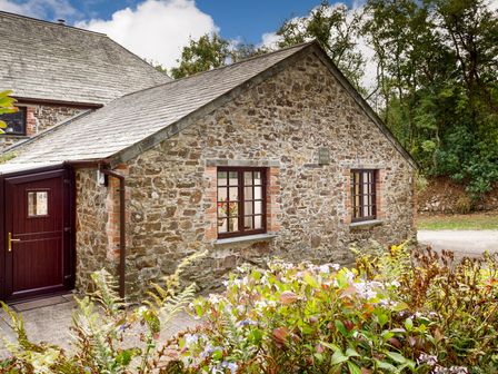 Stone cottage with two windows and a brown door surrounded by plants at The Roundhouse in Warbstow