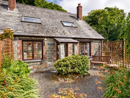 A stone cottage with two windows and a door with a small porch surrounded by plants and a wooden bench at Little Leighs in Warbstow