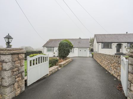 A driveway with open white gates leading to a white house with a garage and a stone wall on the right at Tyddyn Glas in Tyn-y-Gongl near Benllech