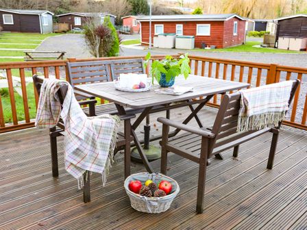 A wooden outdoor deck with a table and chairs with blankets and a basket of apples and pine cones at Eirlys in Caeathro near Caernarfon