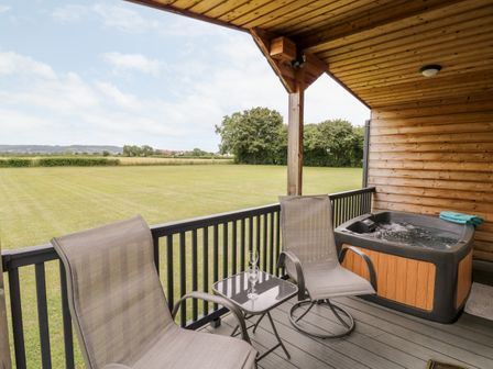 An outdoor covered deck with two chairs a small table with glasses and a hot tub overlooking a grassy field at Cotswold View 1 in Honeybourne