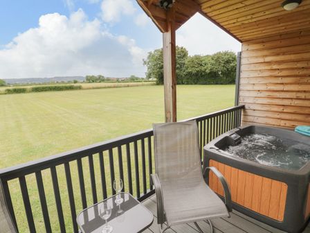 A covered wooden balcony with a hot tub a chair and a small table with two wine glasses overlooking a grassy field at Cotswold View 2 in Honeybourne