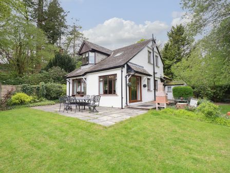 A white two-story house with a patio area and outdoor dining furniture surrounded by green grass and trees at Fir Trees in Bowness-On-Windermere