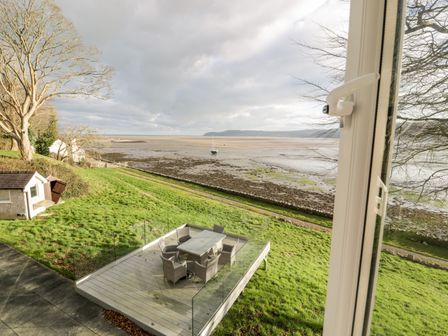 A view of a grassy garden with a raised wooden deck and outdoor dining set overlooking a tidal beach at Porth Llongdy Uchaf in Red Wharf Bay