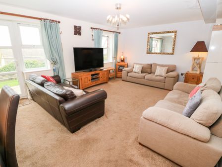A living room with two beige sofas and one black sofa around a TV on a wooden stand at Seascape in Benllech