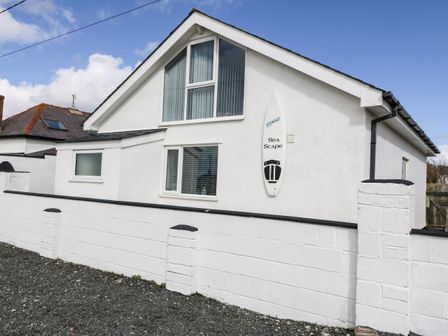 A white house with a surfboard mounted on the wall and a white brick fence in front at Sea Scape in Dinas Dinlle