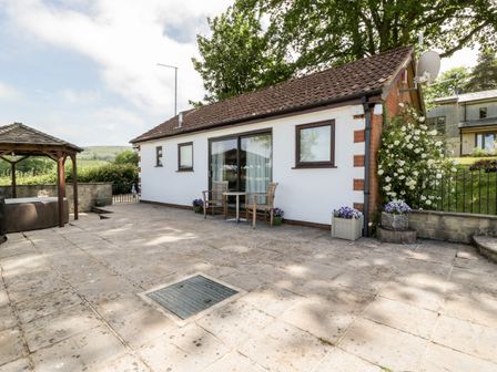 A patio area with stone tiles two wooden chairs and a table next to a small white building with sliding glass doors at The Stables in Crowcombe
