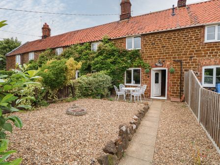 A gravel front garden with a stone border and white patio furniture outside a brick cottage with climbing plants at Tawny Cottage in Heacham
