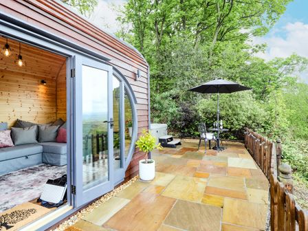 A wooden cabin with glass doors opening to a stone patio with a table umbrella and chairs surrounded by trees at Offas Dyke Escape in St Asaph