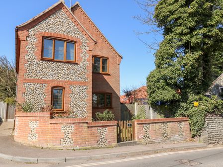 A brick and flint two-story house with wooden windows beside a street and a large tree at Flintstone Cottage in Mundesley