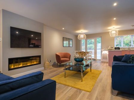 A living room with a wall mounted television above a fireplace a glass coffee table on a yellow rug and a dining area with white chairs at The Neon Rose in Bowness-On-Windermere