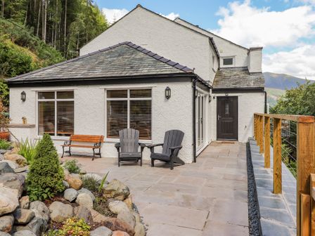 An outdoor patio area with two black chairs a wooden bench stone landscaping and a white house with slate roof at Ladstock Cottage in Thornthwaite near Braithwaite