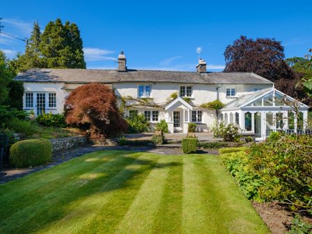 A white cottage with a glass conservatory and a garden with trimmed lawn and shrubs at Orchard Cottage in Outgate near Hawkshead