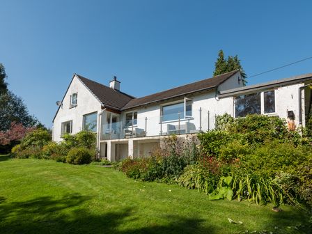 A white house with a glass balcony and garden surrounded by shrubs and grass at Scarsdale in Crosthwaite